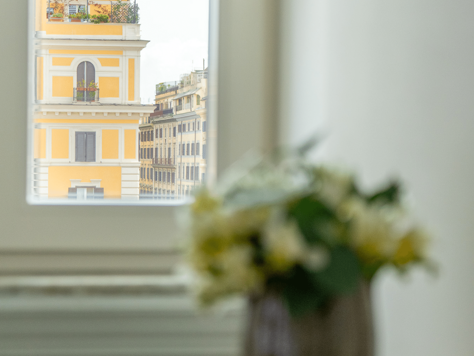 Vue Élégante depuis une Chambre Luxueuse Vue d'un bâtiment historique jaune à travers une fenêtre avec des fleurs floues au premier plan.
